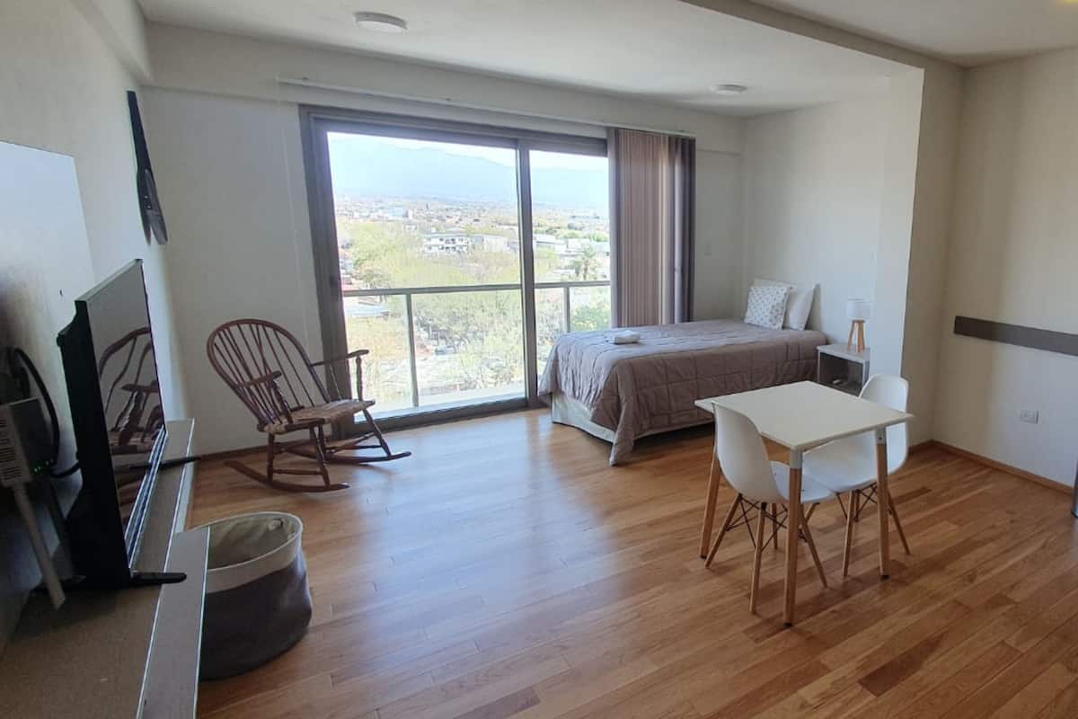 Une chambre spacieuse avec un grand lit, un bureau et une chaise. On y trouve également un fauteuil à bascule et la fenêtre offre une vue sur la ville.
