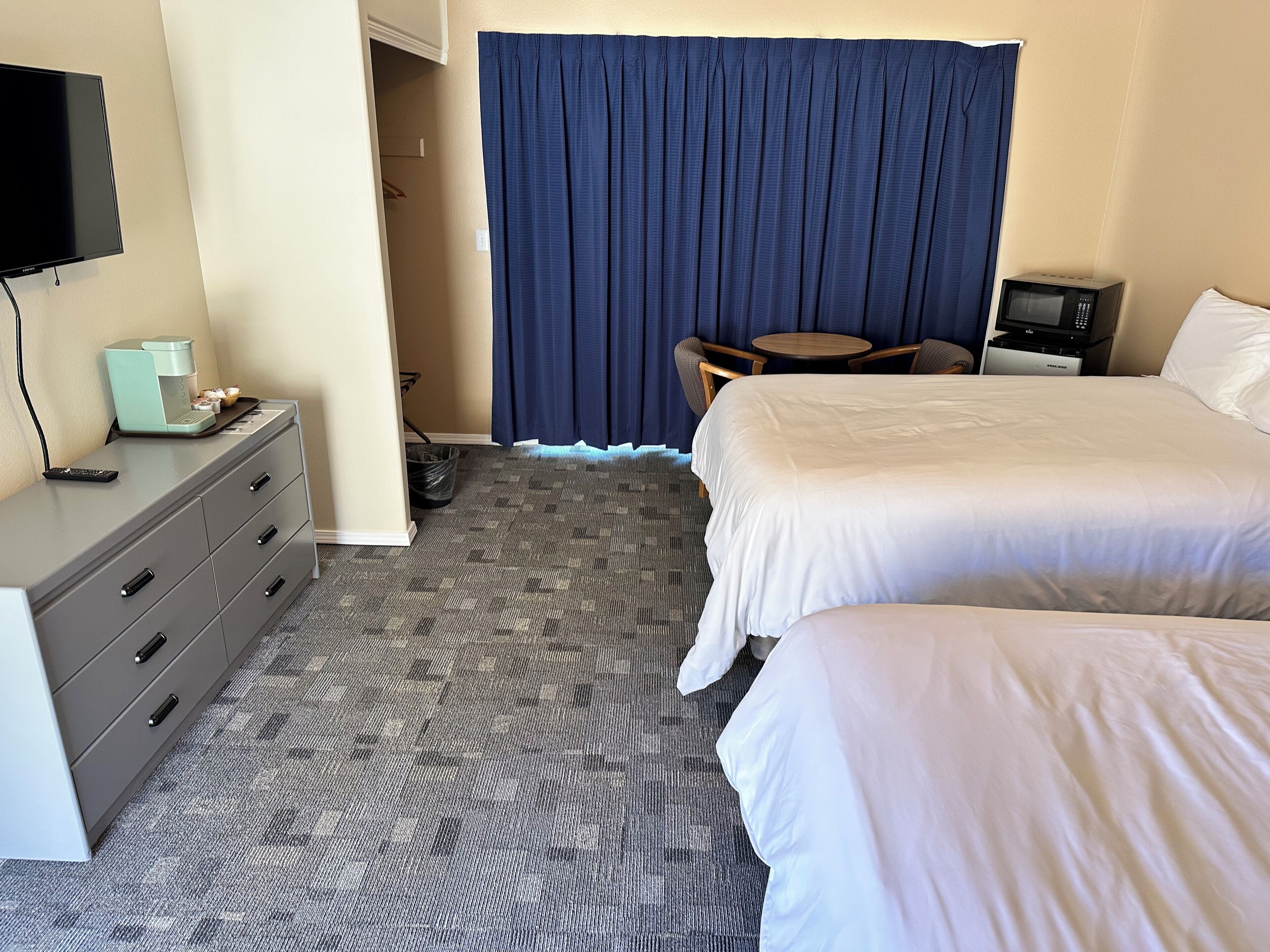 Quarto de hotel com duas camas, uma mesa de escritório com cafeteira, televisão, micro-ondas e uma pequena mesa com cadeiras.