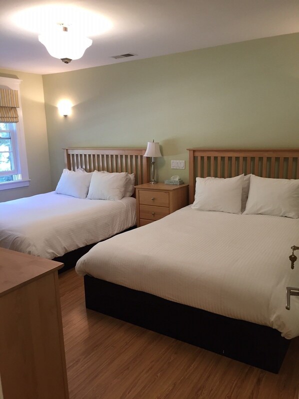 A hotel room with two single beds, a bedside table with a lamp, a small drawer, and a wooden desk.