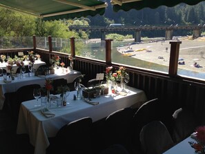 A restaurant with tables set for dining, overlooking a river with boats and a bridge in the background. - Highland Dell Lodge (Monte Rio)