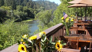 Une terrasse en bois offrant une vue sur une rivière et une forêt. On y trouve une table avec un vase rempli de tournesols et un drapeau américain.