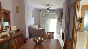 A dining area with a wooden table, chairs, a bowl, and a lamp. There is a sofa and a window with curtains. - Chiclana sunset (Chiclana de la Frontera)