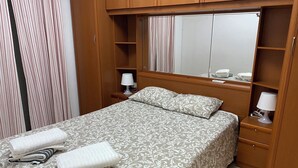 A neatly made bed with a patterned bedspread and pillows, a bedside table with a lamp, a mirror above the bed, and a wooden wardrobe with a mirror. - Chiclana sunset (Chiclana de la Frontera)