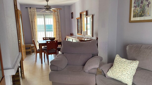 A living room with a grey sofa, a wooden coffee table, a dining area with a table and chairs, a ceiling fan, and a window with curtains. - Chiclana sunset (Chiclana de la Frontera)