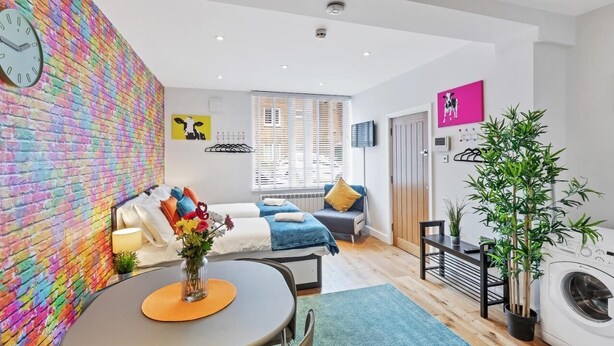 A modern living room with a colorful brick wall, a sofa, a dining table with a vase of flowers, and a washing machine.