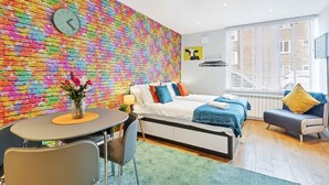 A modern room with a colorful brick wall, a bed with blue and orange pillows, a small table with a chair, and a window with blinds.