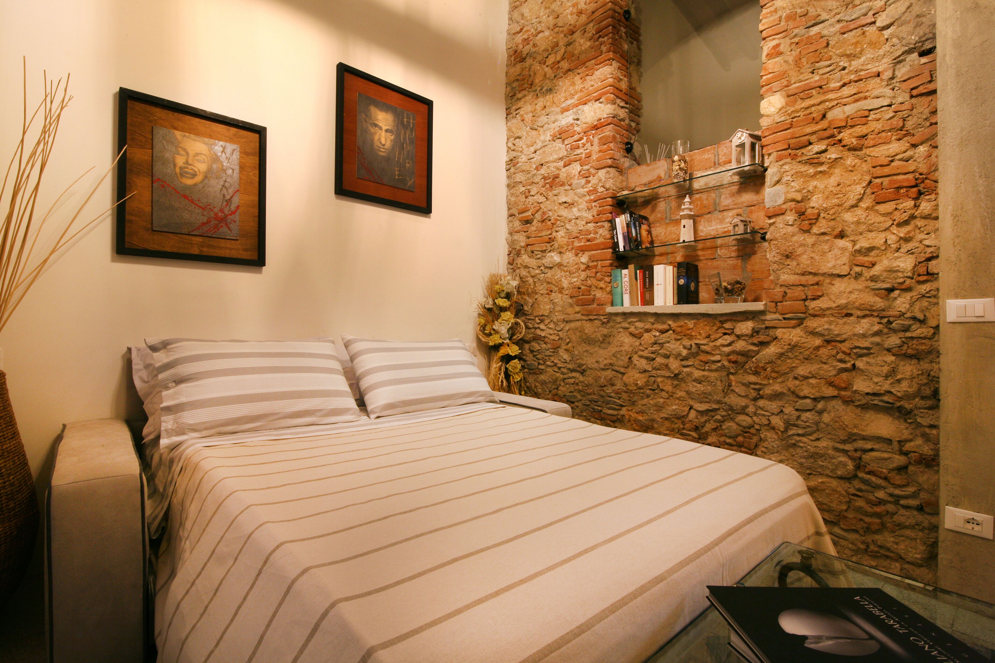 A bed with striped bedding, a stone wall, and a shelf with books and decorative items.