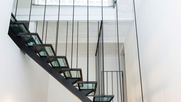Un escalier moderne avec des rampes en verre et des éléments décoratifs en métal noir, menant à un plafond haut surmonté d’une verrière.