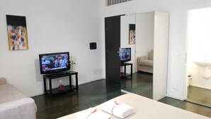 Une chambre d’hôtel moderne avec un grand miroir, un téléviseur posé sur un meuble, un canapé beige et une petite table sur laquelle se trouve un vase.