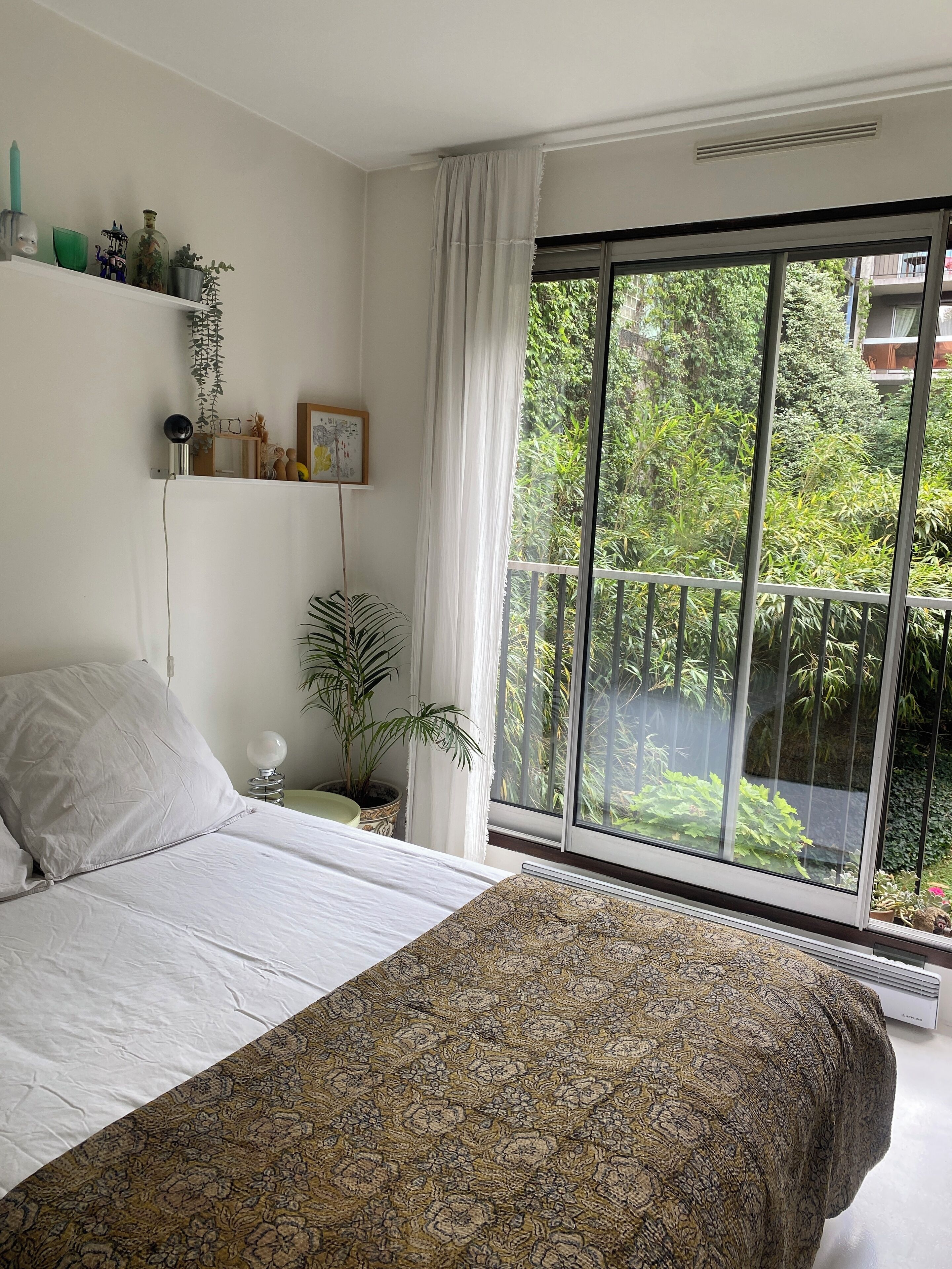 Une chambre avec un grand lit, une fenêtre donnant sur un paysage verdoyant, une étagère avec des objets décoratifs et une table de chevet avec une lampe.