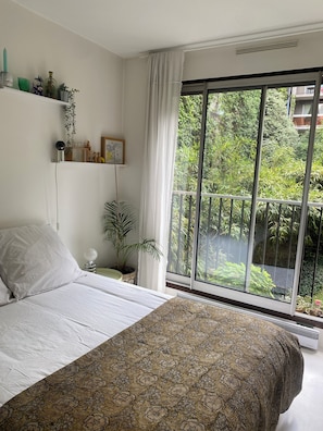 Um quarto com uma cama grande, uma janela com vista para vegetação, uma prateleira com objetos decorativos e uma mesa de cabeceira com abajur.