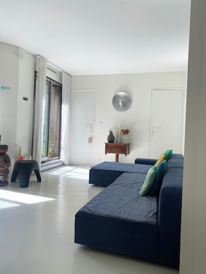Uma sala de estar moderna com um sofá azul escuro, uma pequena mesa lateral de madeira e uma grande janela com cortinas.