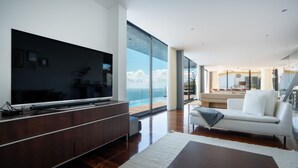 Un salon moderne avec un grand téléviseur à écran plat posé sur une console en bois, un canapé blanc et une vue sur l’océan grâce à de grandes fenêtres du sol au plafond.