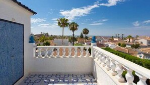 Een balkon met een patroonvloer, een witte balustrade en uitzicht op een woongebied met palmbomen en huizen onder een heldere lucht.