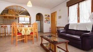 Een woonkamer met een donker leren bank, een houten eettafel met stoelen, een glazen salontafel, en een keuken met een rondbog venster.