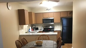 A dining area with a table set for six, surrounded by wooden chairs. A microwave and stove are visible in the kitchen area behind the table.