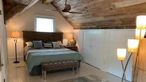 Quarto com teto de madeira, cama com colcha verde, duas luminárias de cabeceira, um banquinho e uma janela grande.