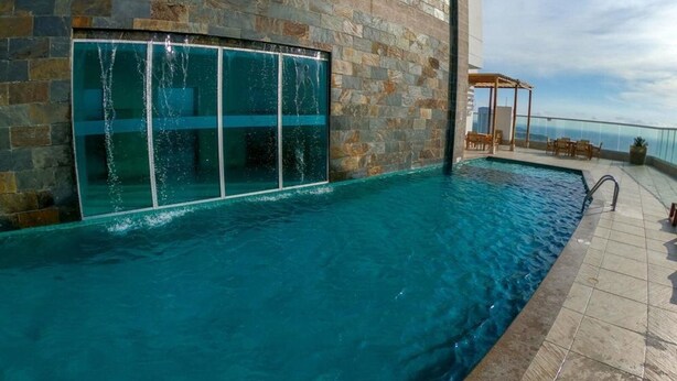 Ein Swimmingpool mit einer Steinwand und einer Glastür, umgeben von einer gefliesten Terrasse und Gartenmöbeln.