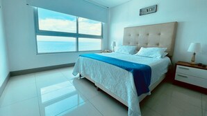 Ein Schlafzimmer mit großem Fenster und Meerblick, einem Bett mit blauer Bettdecke, einem Nachttisch mit Lampe und einem Schild mit der Aufschrift 'BEACH'.