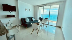 Ein modernes Wohnzimmer mit einem dunkelgrünen Sofa, weißen Stühlen, einem flachbildfernseher an der Wand und einer großen Glasschiebetür, die auf einen Balkon mit Meerblick führt.
