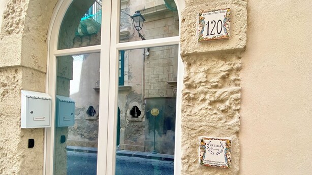 Una porta bianca a due ante con pannelli di vetro, incastonata in un arco, con un muro in pietra e una targa con il numero civico.