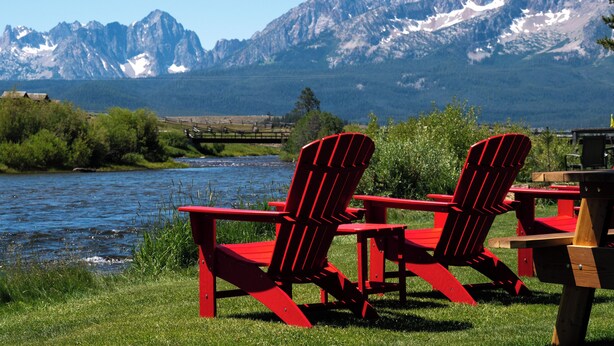 Dos sillas Adirondack rojas junto a un río, con montañas al fondo.
