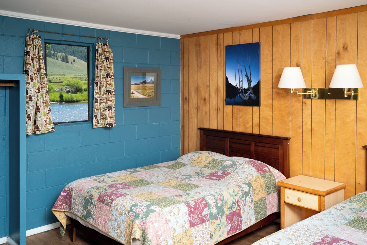 A bedroom with a wooden headboard, a bed with a floral quilt, a bedside table with a lamp, and a window with curtains.