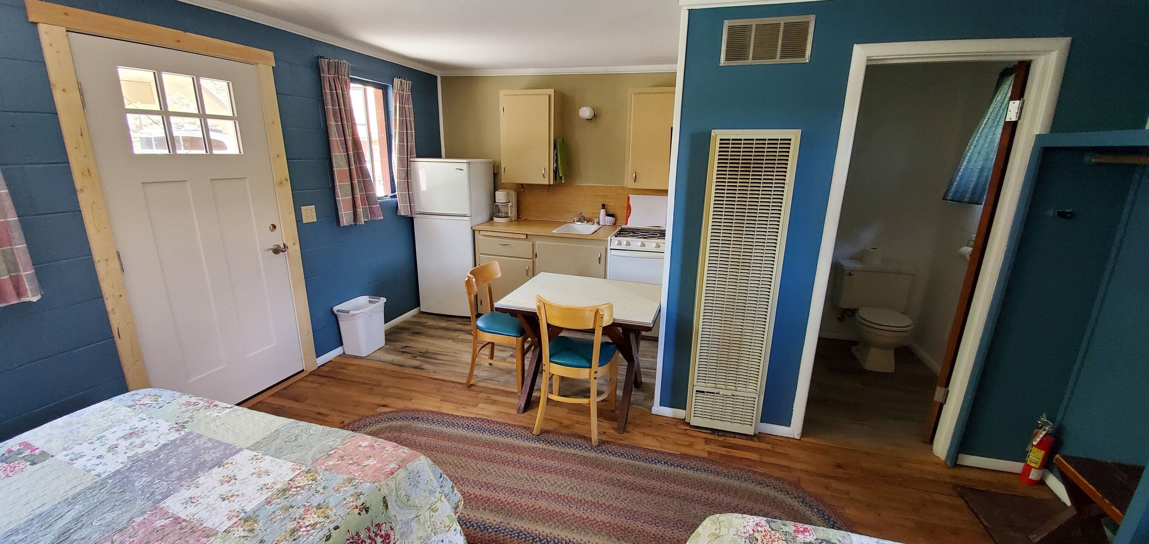 A compact room with a bed, a small dining table with chairs, a kitchenette with a stove, and a toilet.