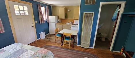 A compact room with a bed, a small dining table with chairs, a kitchenette with a stove, and a toilet.