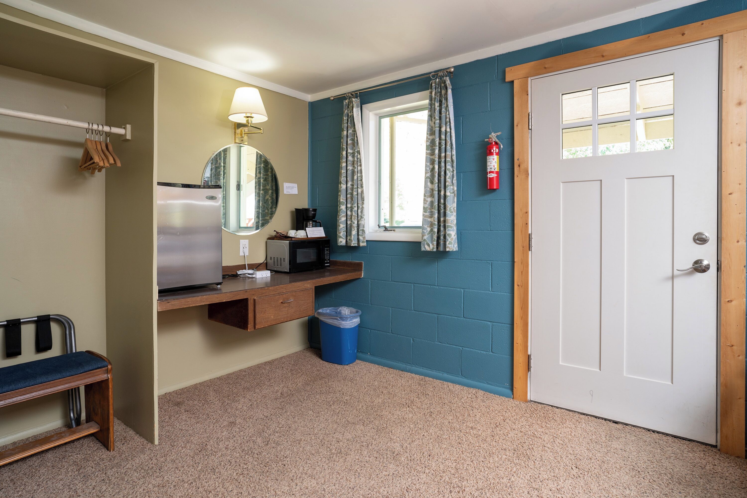 A compact room with a microwave, a small table, a bench, a mirror, a window with curtains, and a door.