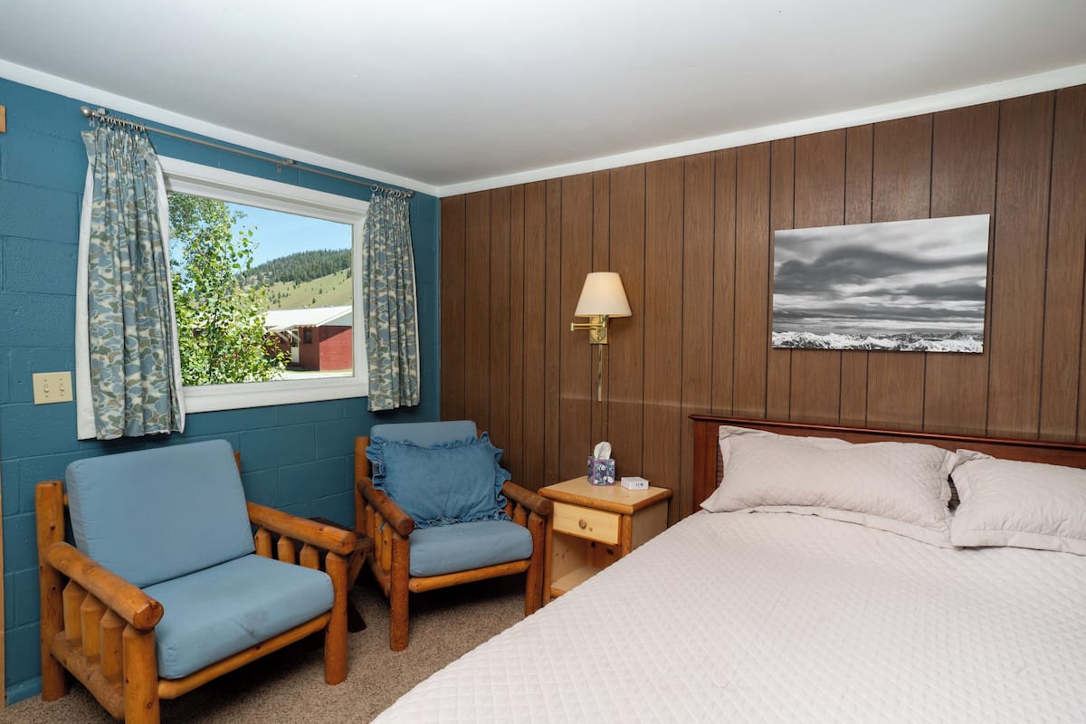A bedroom with a bed, two wooden chairs, a nightstand, a lamp, and a window with curtains.