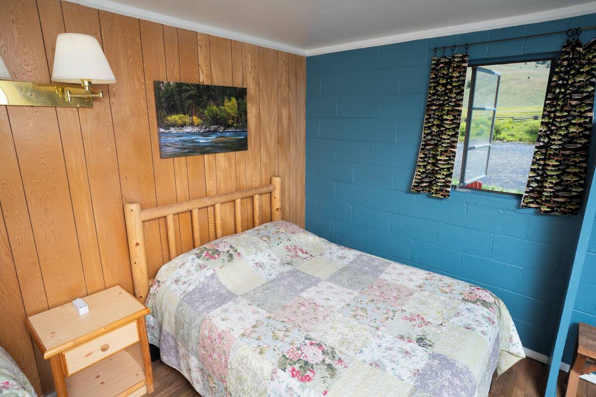 A bedroom with a wooden headboard, a floral quilt, a nightstand, a wall-mounted lamp, a painting of a river, and a window with curtains.