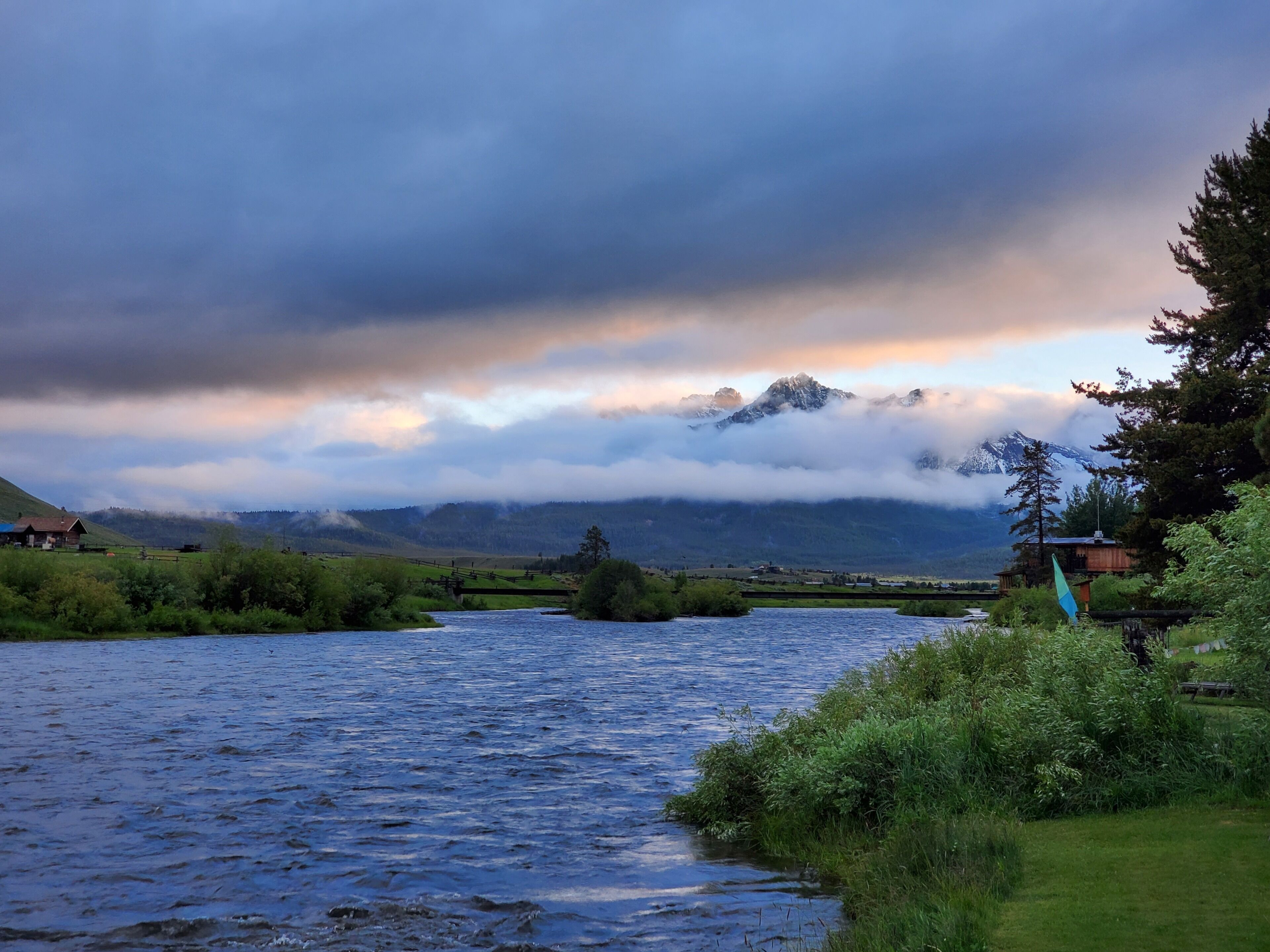 Un río serpentea por un paisaje rural con casas a orillas, rodeado de vegetación y montañas al fondo.