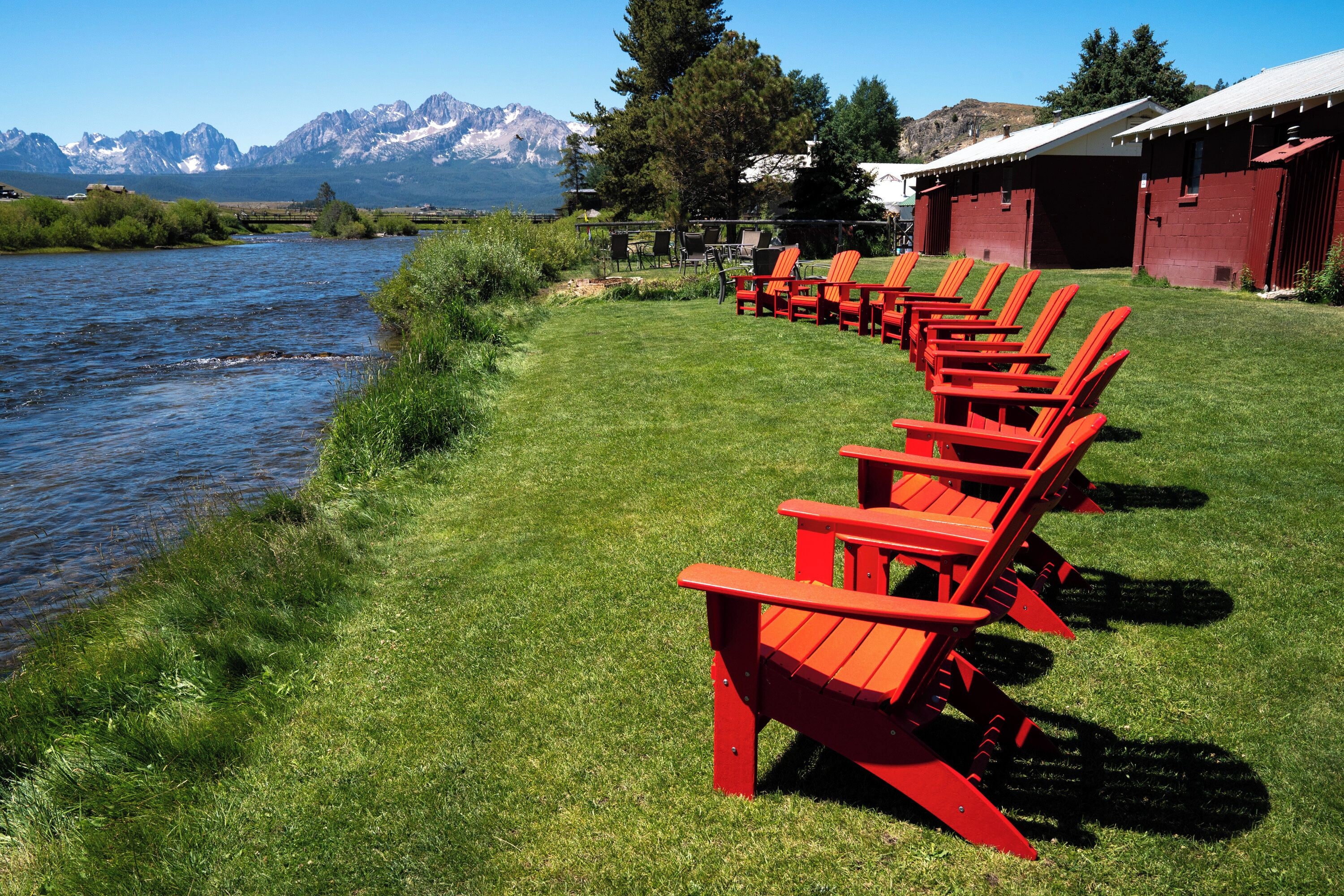 A riverside view with red Adirondack chairs lined up on the grass, a red barn, and mountains in the background.