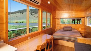 Una cabaña de madera con una cama, un escritorio y ventanas amplias que ofrecen vistas a un lago y colinas.
