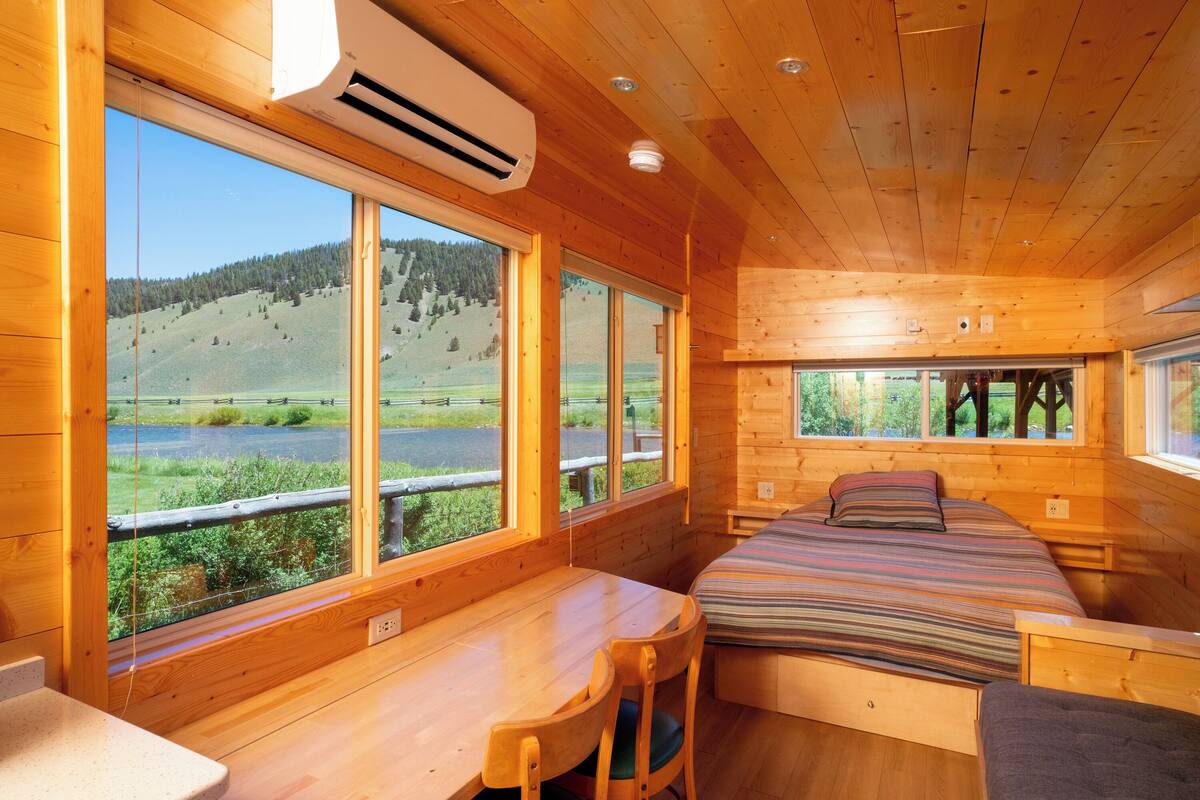 A wooden cabin with a bed, a desk, and large windows offering a view of a lake and hills.