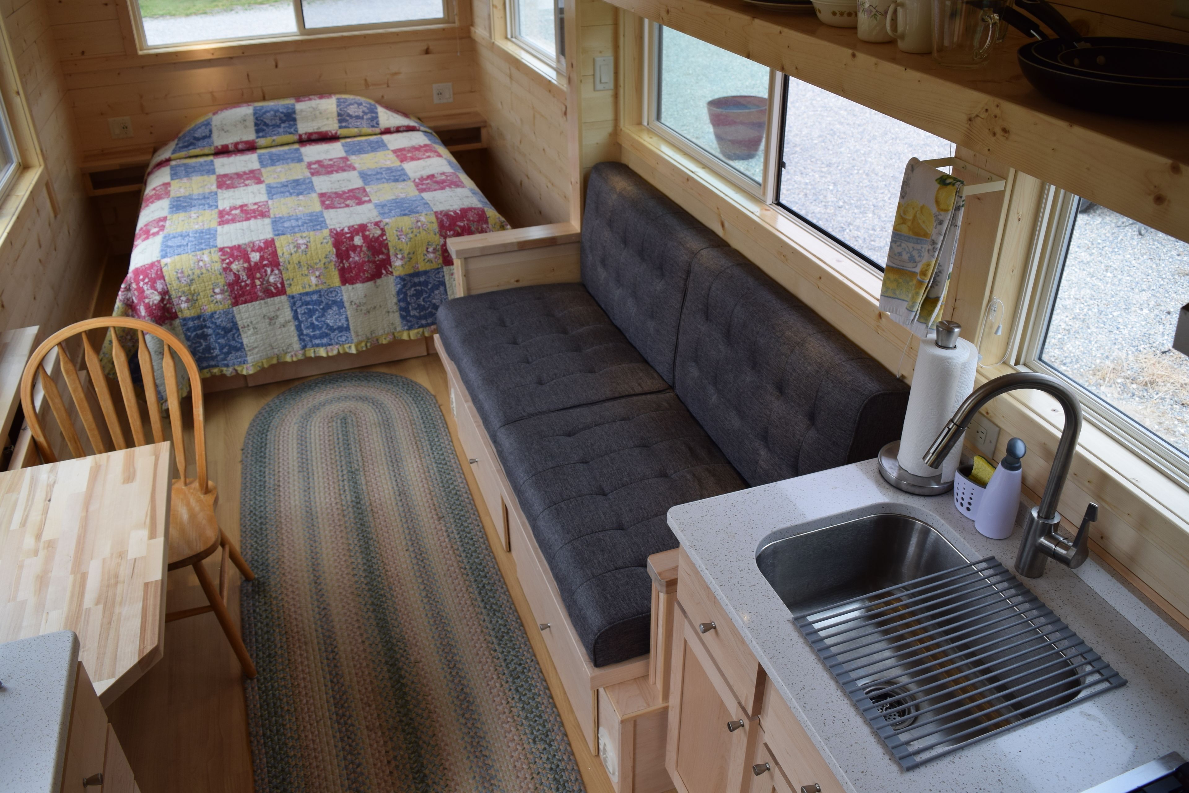 A compact cabin with a kitchenette, a bed with a colorful quilt, a gray sofa, and a window overlooking a gravel path.