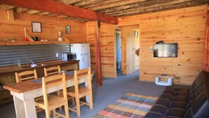 A cozy cabin interior with a wooden dining table, chairs, a sofa, a television, and a kitchen area with a refrigerator and microwave. - mandalas demar (Punta del Diablo)