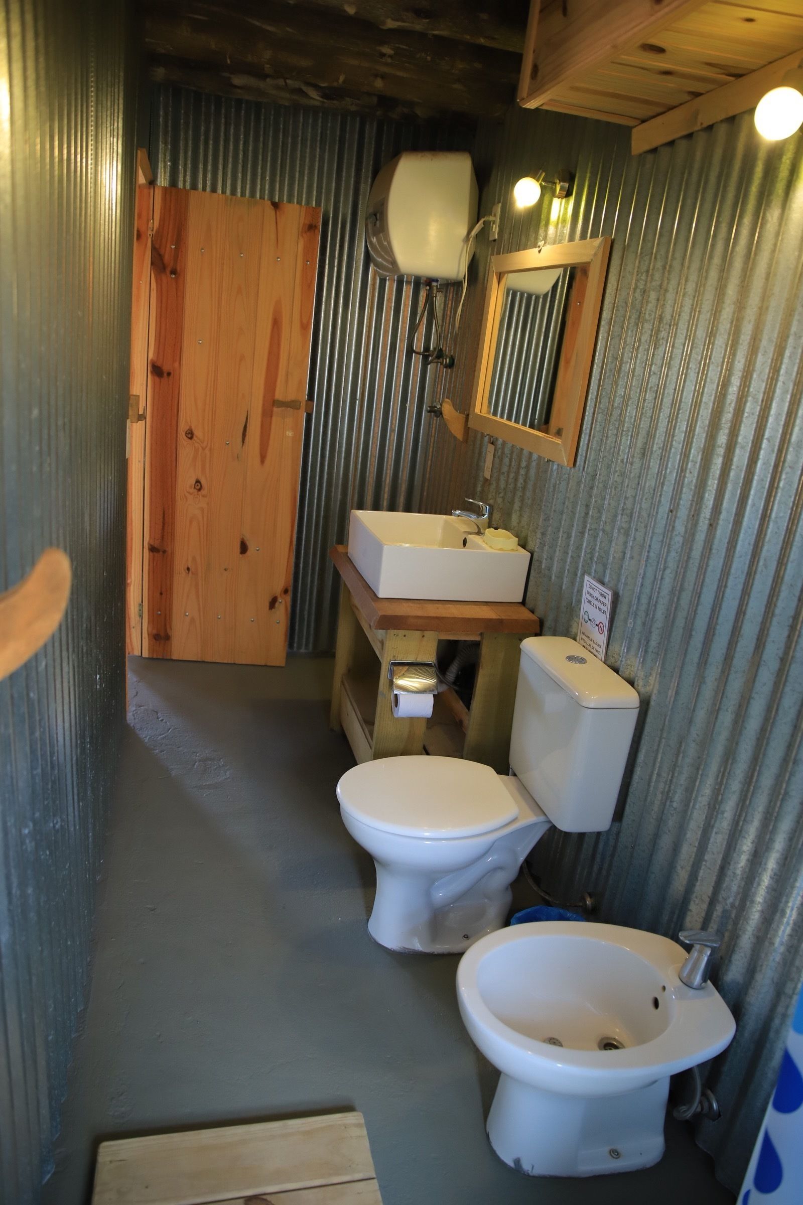 A compact bathroom with a wooden door, a white toilet, a bidet, and a sink with a mirror above it.
