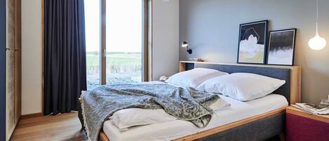 A bedroom with a large bed, a wooden floor, a window with curtains, and two framed pictures on the wall.
