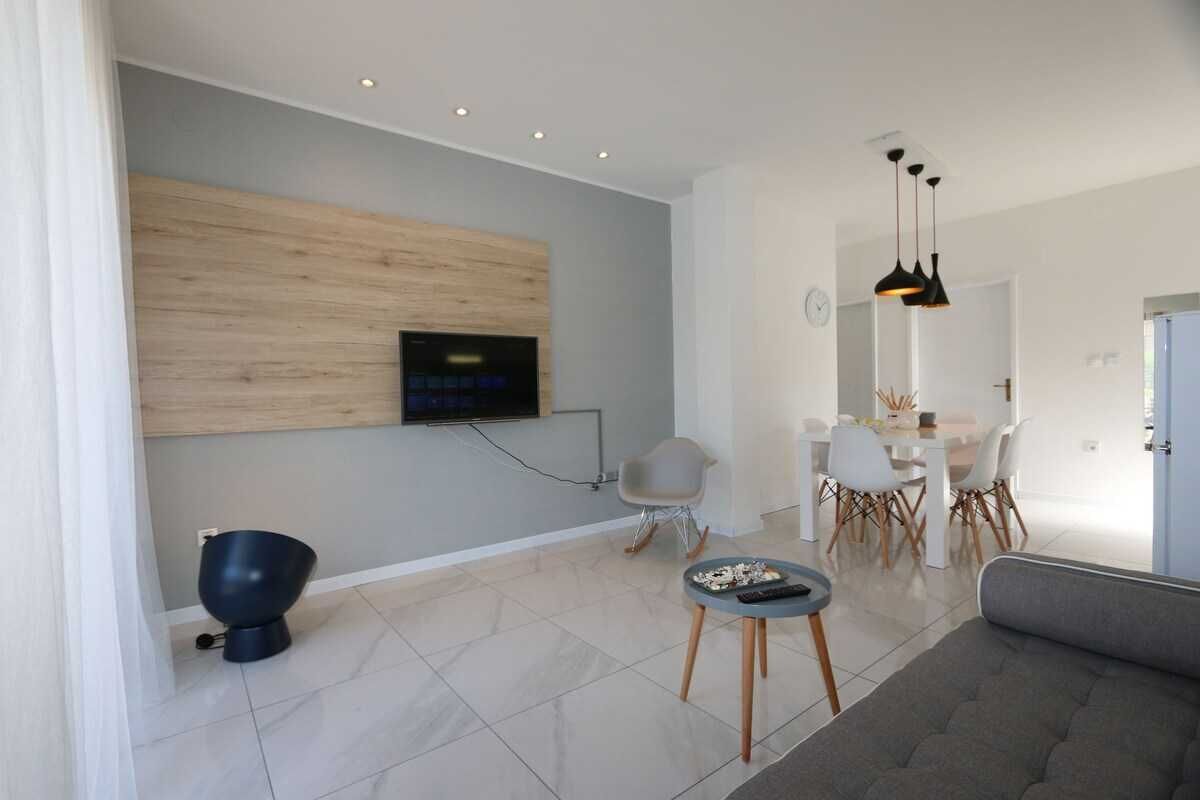 A modern living room with a flat-screen TV mounted on a wooden panel, a dining area with a white table and chairs, and a grey sofa.