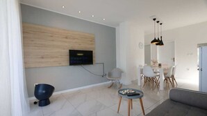 Una sala de estar moderna con un televisor de pantalla plana montado en un panel de madera, un comedor con una mesa y sillas blancas, y un sofá gris.