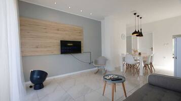 A modern living room with a flat-screen TV mounted on a wooden panel, a dining area with a white table and chairs, and a grey sofa.