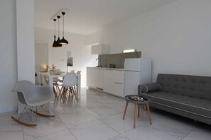 Uma cozinha moderna com armários brancos, uma área de jantar com mesa e cadeiras brancas, um sofá cinza e uma pequena mesa lateral.