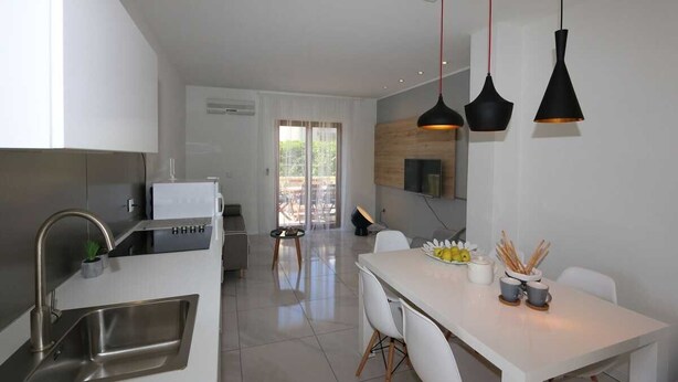 Una cocina moderna con un lavamanos de acero inoxidable, una zona de comedor con una mesa y sillas blancas, e iluminación colgante.