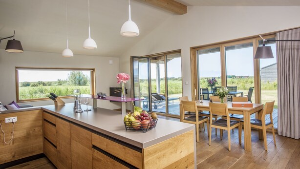Una cocina moderna con armarios de madera, una isla central con fruta, luces colgantes y un comedor con sillas y una mesa de madera.