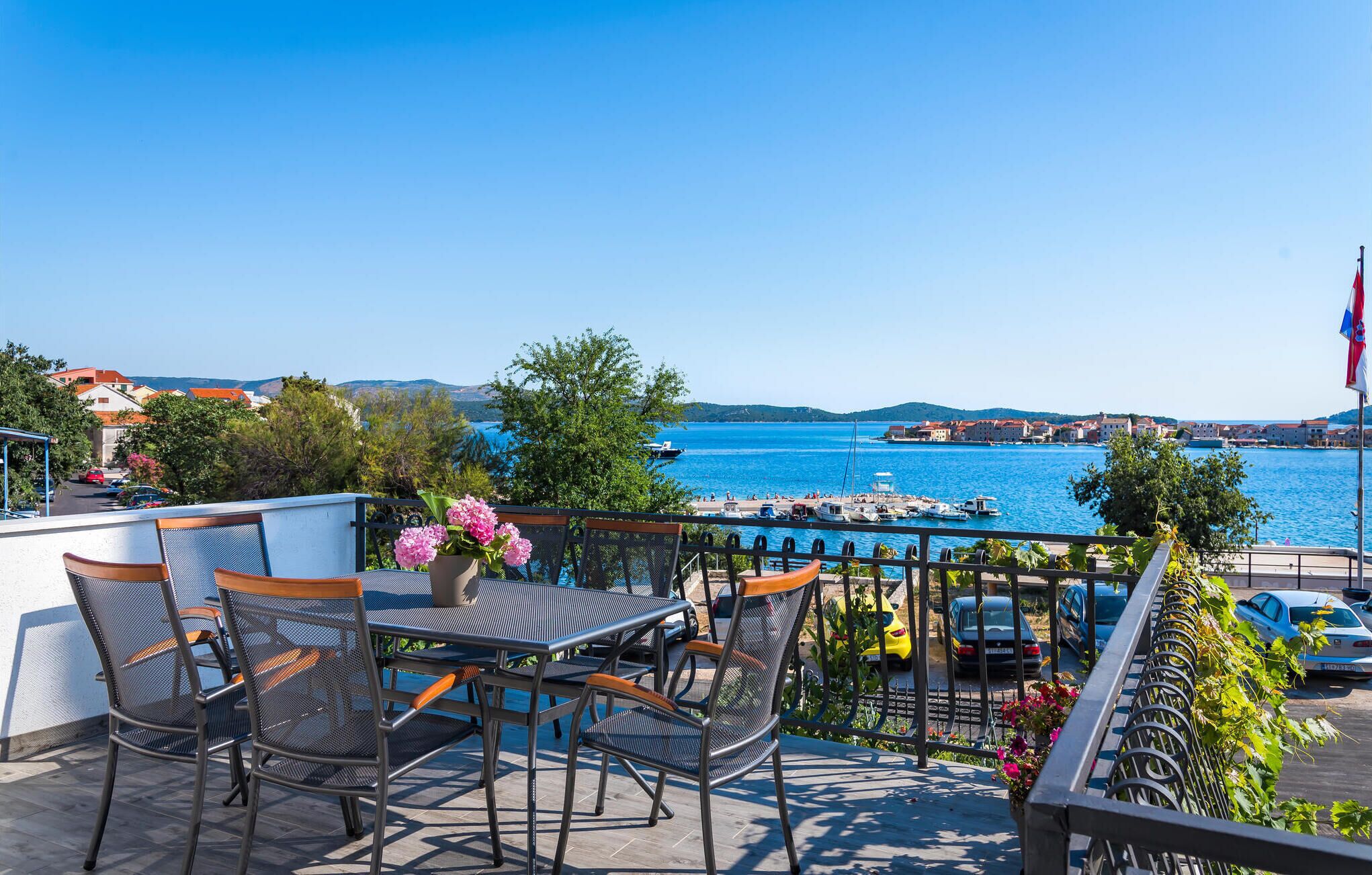 Ein Balkon mit Tisch und Stühlen, mit Blick auf einen Yachthafen und ein tiefblaues Meer.
