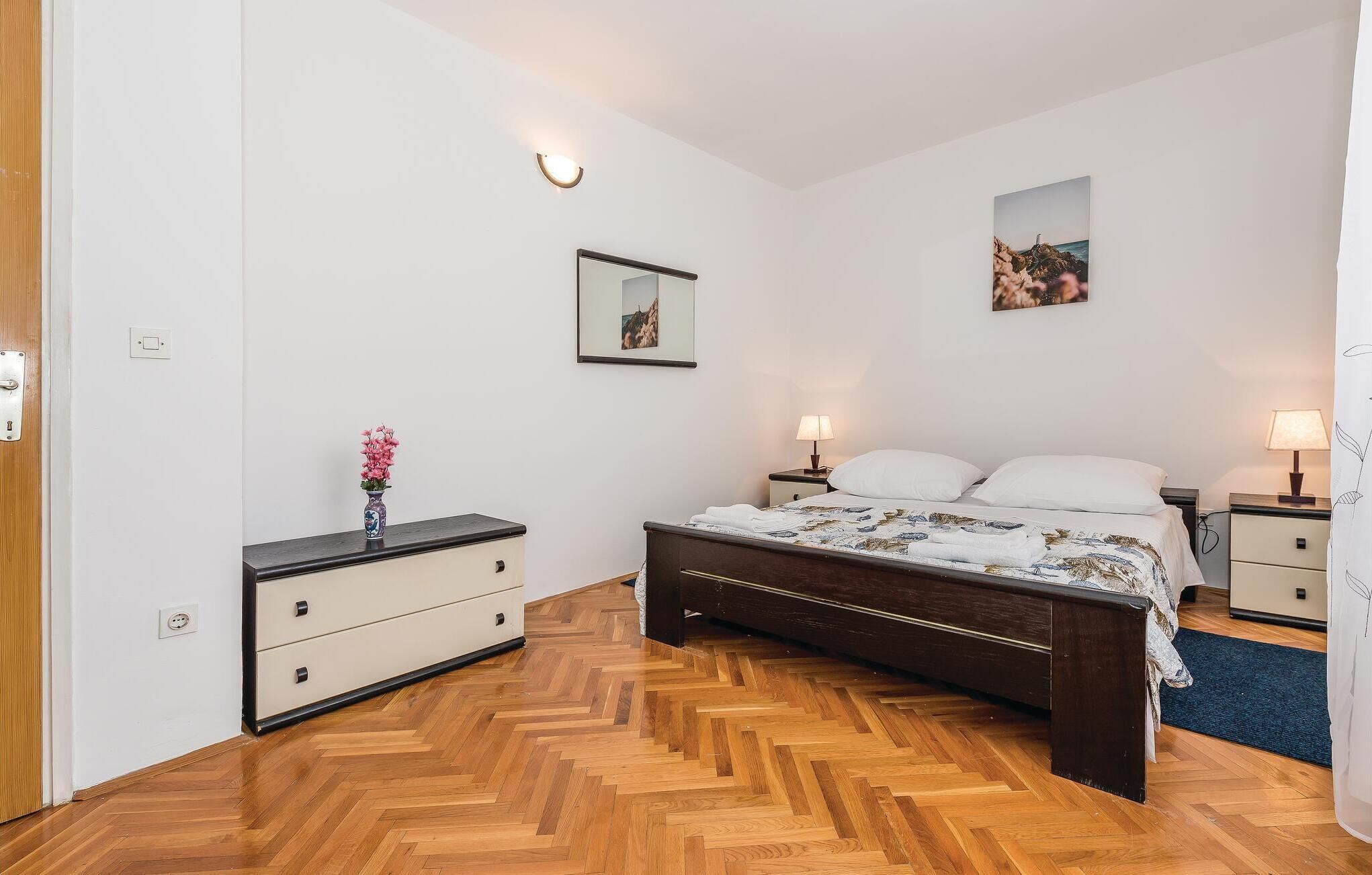 A bedroom with a wooden herringbone floor, a bed with white linens, two bedside tables with lamps, a nightstand, a dresser, and a wall-mounted light fixture.