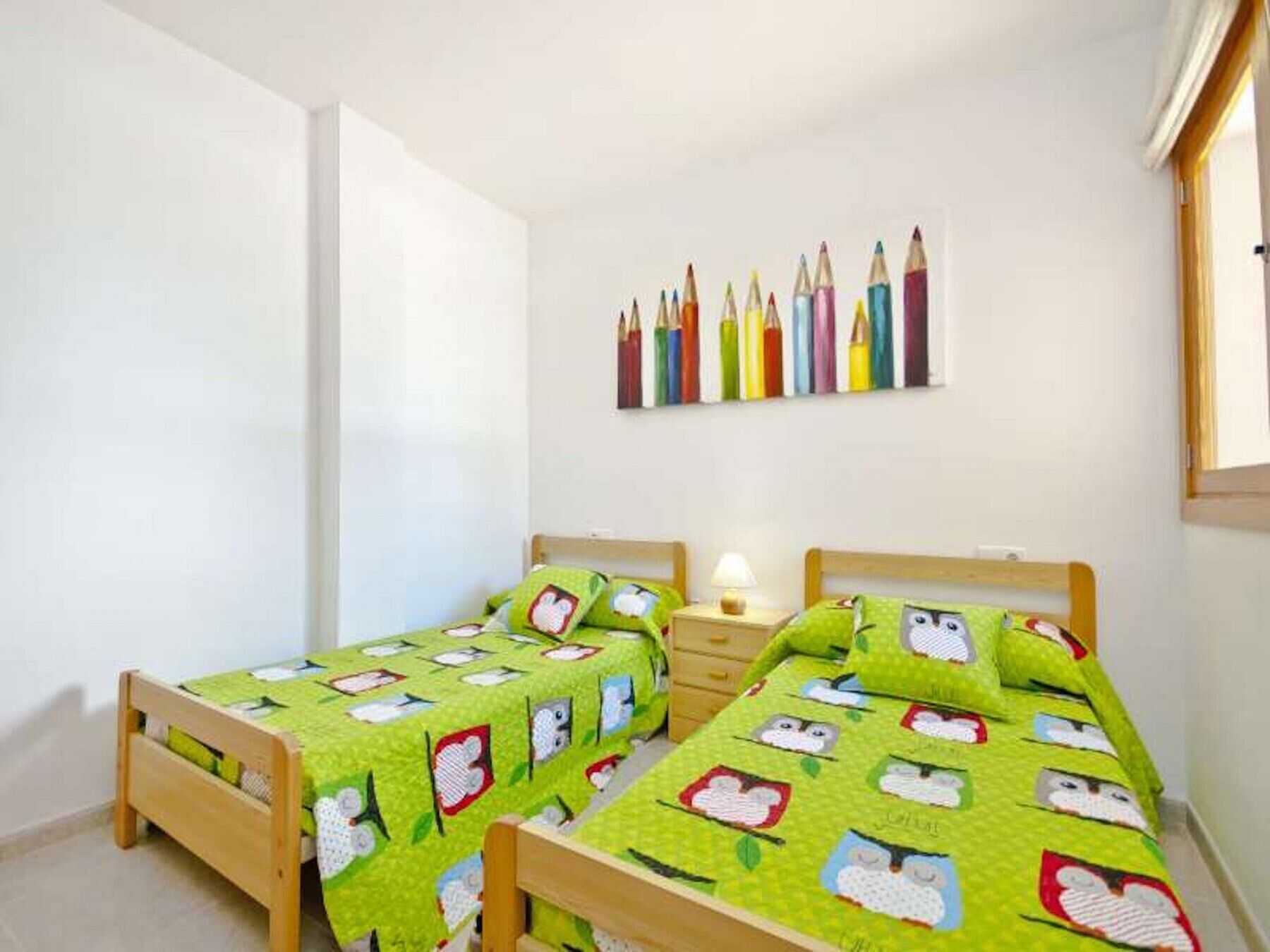 A bedroom with two wooden beds, each with a green quilt featuring owl patterns. There is a wall-mounted shelf with colorful pencils above the beds.