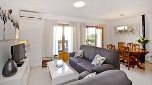 En moderne stue med en grå sofa, et sofabord, et fjernsyn og et spiseområde med træstole og et bord.